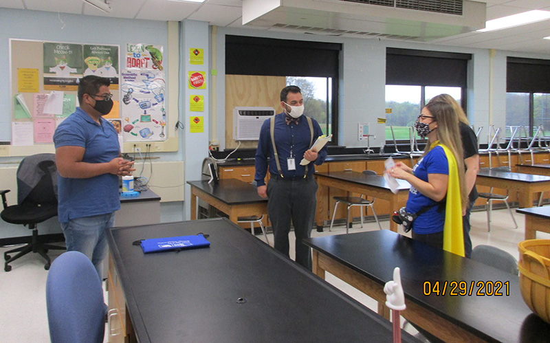 A man by a long lab counter stands and watches as a woman in a blue shirt and gold cape presents him with a key chain. Two others are in the room watching. All are wearing masks.