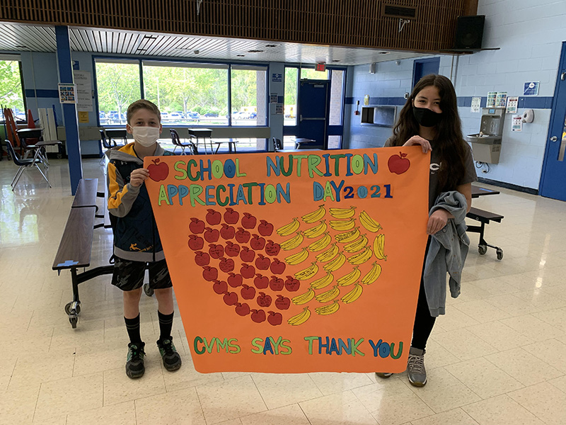 Two middle school students - a boy on the left and a girl on the right, hold a large orange sign that has a  heart made from material in the shape of apples and bananas. It says School Nutrition Appreciation Day 2021 CVMS  thanks you.