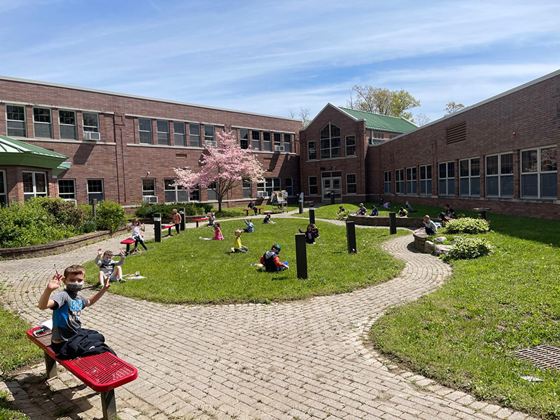 A wide view of a courtyard surrounded by a brick building. It has a circular path with a patch of grass in the midde. All around it are elementary students, at a safe distance apart from each other, sitting on the ground and painting.