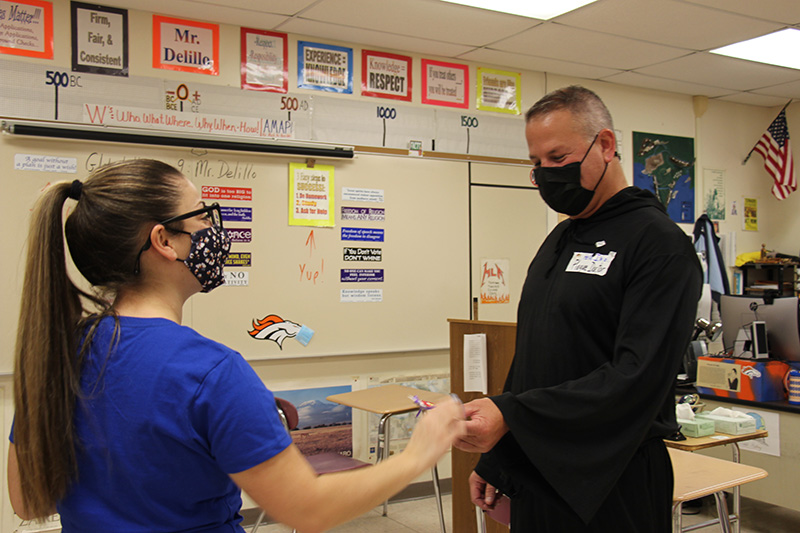 A woman in a blue shirt hands a key chain to a man all dressed in black. Both are wearing masks.