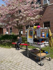 A cart filled with art supplies on the right. Just in front of it is a student sitting on a red bench painting. There is a beautiful tree with pink blossoms on it.