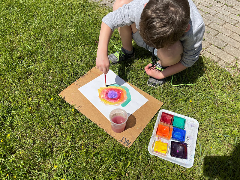 An elementary student squats down in the grass and paints with many different colors.