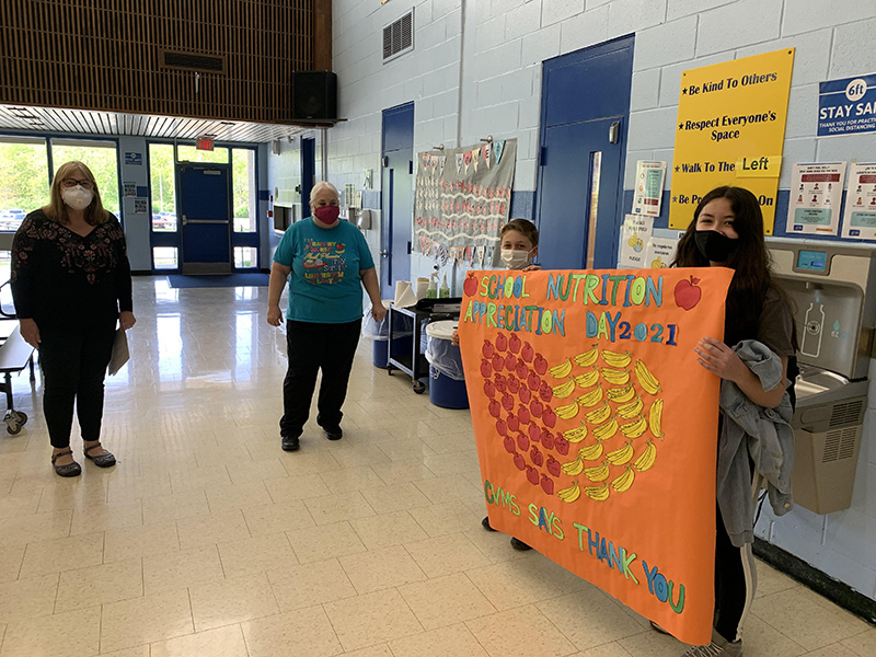 Two middle school students - a boy on the left and a girl on the right, hold a large orange sign that has a  heart made from material in the shape of apples and bananas. It says School Nutrition Appreciation Day 2021 CVMS  thanks you. Two women are standing looking at the sign. 