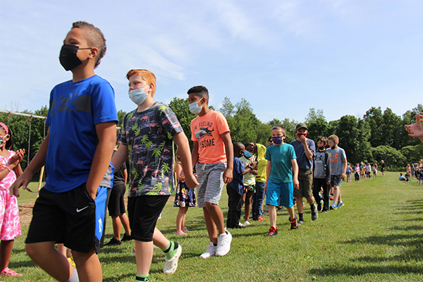 A line of fifth-grade kids walk on grass, with a beautiful blue sky above. On either side of them are kids clapping.