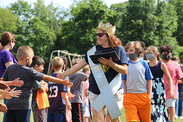 A parade of fifth grade kids follow a woman who is wearing a big gold crown nwith silver stars on it and a sash. They are high fiving other kids that have lined a path for them.