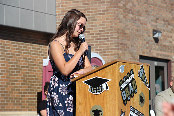 A young woman in a blue and white dress stands at a wooden podium. Her hair is long and brown. She is talking into a microphone. The podium has graduation decorations on it!