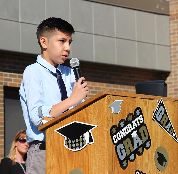 An 8th grade young man wearing a light blue shirt and tie, standing at a podium  holding a microphone. On the podium are graduation decorations.