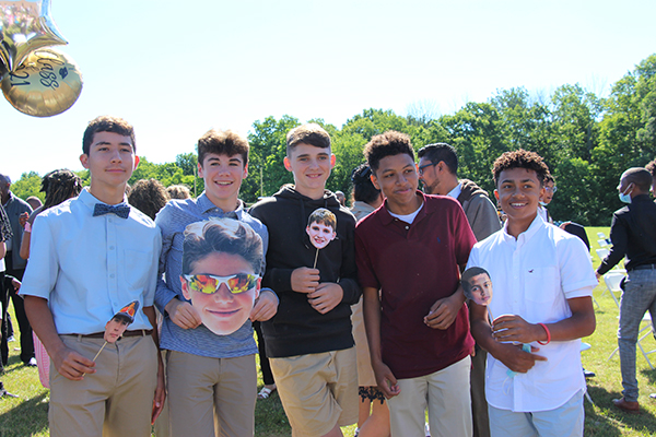 A group of five eighth grade boys stand together smiling. They are holding cardboard cut outs of their own picture.