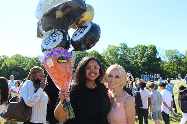 Two girls in eighth grade stand together smiling. The girl on the left is wearing a black dress and holding lots of black, white and gold graduation balloons. She has a bouquet of flowers too. The girl on the right is blonde wearing a light colored dress. They are both smiling.