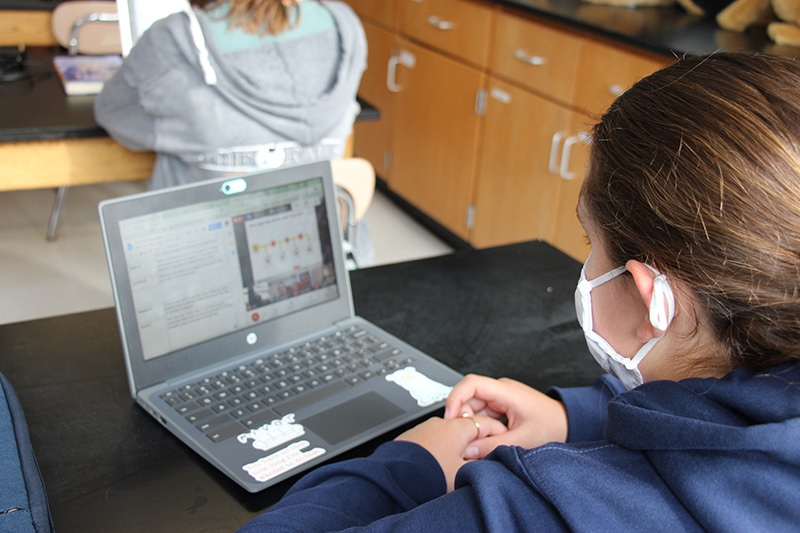 A student wearing a white mask and a blue shirt sits at the desk looking at a Chromebook. The photo is taken over the student's shoulder.