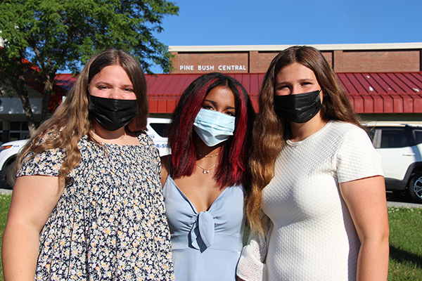 Three eighth-grade girls stand together with arms around each other. Girl on left is wearing a black and white print shirt and black mask. She has long blonde hair. The girl in center is wearing a blue dress and has dark hair with red streaks. The girl on the right has long blonde hair and is wearing a white dress. behind them is a red building, a tree and blue sky. They are all wearing masks.