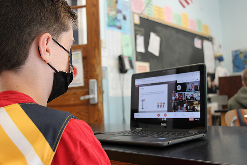 A male student wearing a black mask and a yellow and black shirt with red sleeves watches a presentation on his Chromebook. The photo is taken over his shoulder so you can see the screen. 
