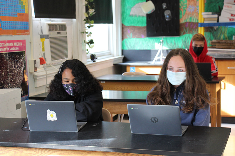 Two girls wearing masks, each with long dark hair, watch a presentation on their Chromebooks in a classroom.
