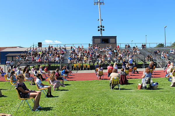 On an athletic field, people inthe stands and students sitting in yellow chairs on the field, socially distanced.