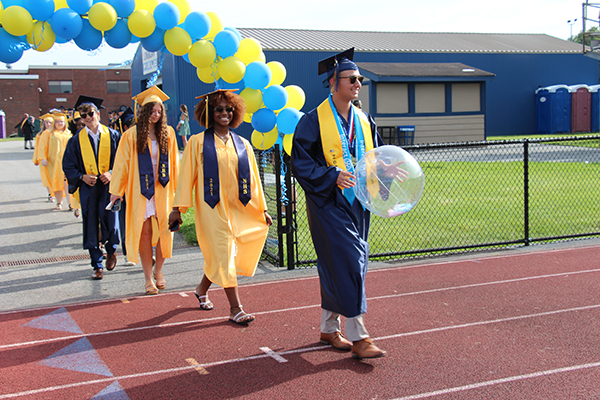 A group of high school seniors dressed in blue caps and gowns and gold caps and gowns walk onto a track under an arch of blue and gold balloons.