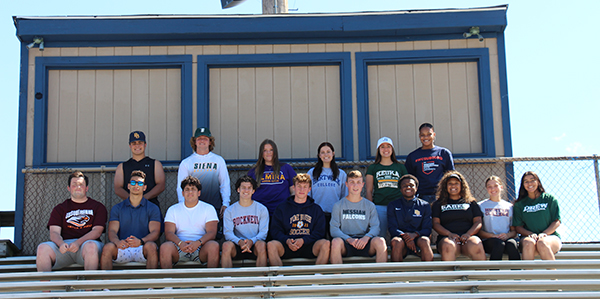 Sixteen high school seniors are sitting on bleachers in two rows. Blue sky in the background and a press box behind them.