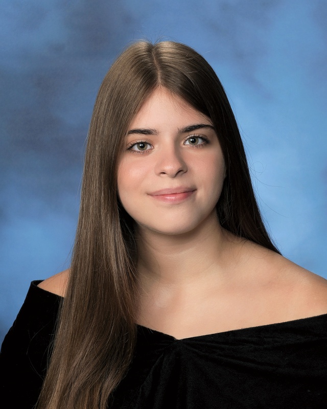 A young woman in aformal high school graduation photo. She has long brown hair, parted in the middle, and is wearing a black off-the-shoulder sheath.