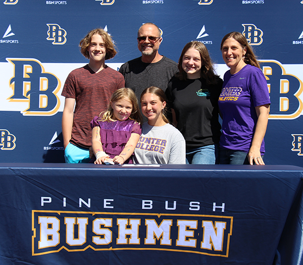 Four adults stand behind a young woman and a little girl who are sitting at a table. The table is covered in a blue cloth that says Pine Bush Bushmen. The background also is blue with PBs on it.