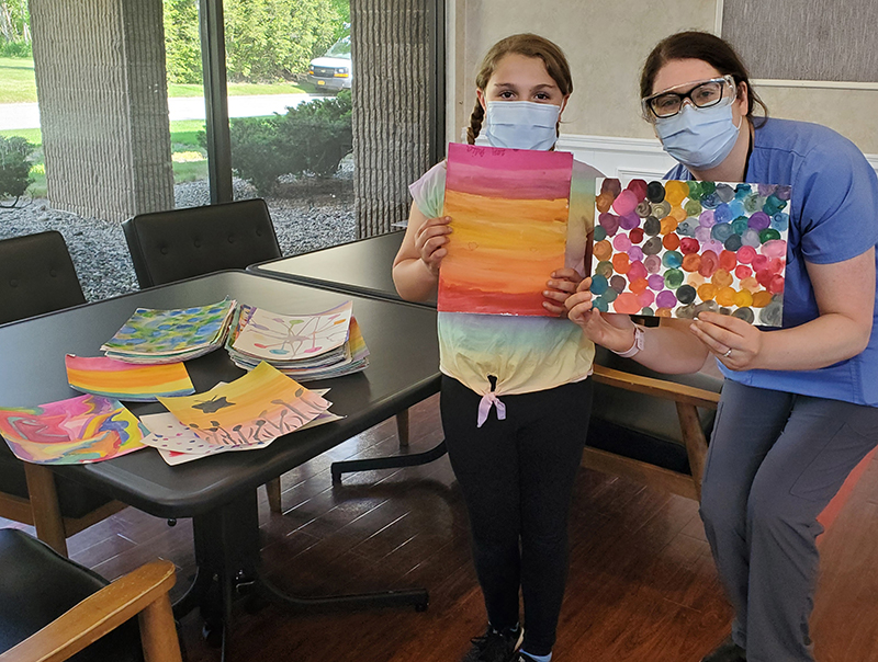 A young girl with her hair pulled back holds a piece of paper that is painted with several different colors. NExt to her is a woman with glasses also holding a piece of paper that is painted. Both are wearing masks. There is artwork on the table beside the girl.