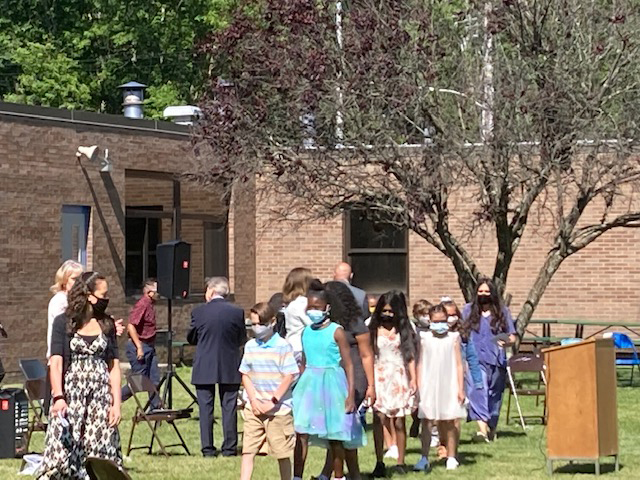 A brick building and a tree are in the background as elementary students walk on grass in a line.