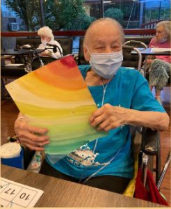 An older man wearing a blue shirt and paper mask holds up a painting that looks like a rainbow of colors.