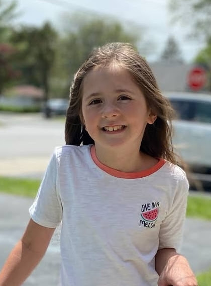 A little girl with long brown hair smiling. She is wearing a white t-shirt with orange trim around the neck. On the side it says One in a Melon with a picture of a watermelon on it.