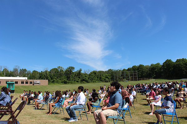 Students sitting in blue chairs out on a field of grass with a large number os parents in chairs behind them. Beautiful blue sky with a long whispy white cloud above.