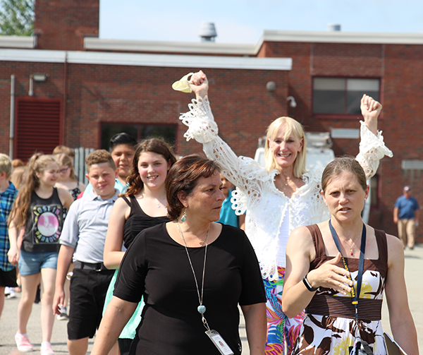 A group of fifth-grade students and a few teachers march onto a field.  One of the teachers, who has blonde hair and is wearing a white shirt, has here arms up in the air and is smiling.