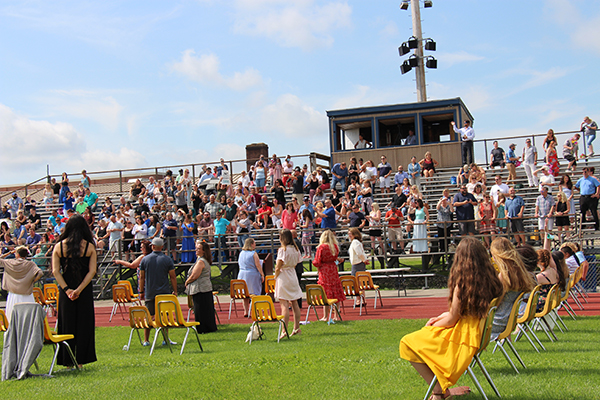 A view from an athletic field. There are fifth-grade students sitting in chairs on the grass and a large group of parents in the stands beyond them. The sky is blue with some white clouds.