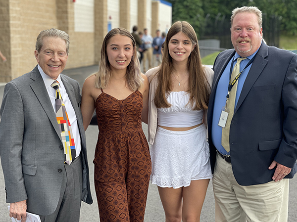 Two adult men on either side of the group. Two high school seniors are in the center. The men have suits on . The young woman center left has a brown print jumpsuit on. She has shoulder length blonde hair. The young women center right has long brown hair and is wearing a white shirt and shirt. All are smiling.