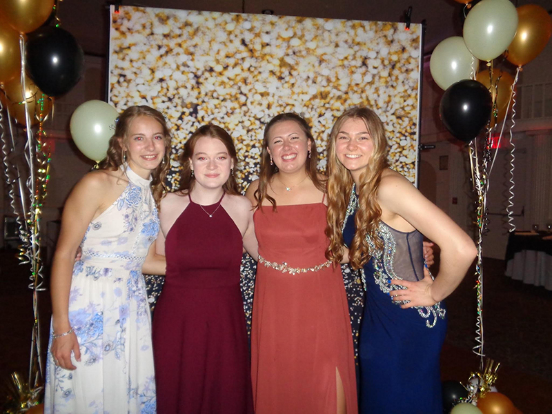 Four young women all dressed in long gowns, smile for the camera in front of a sparkly background.