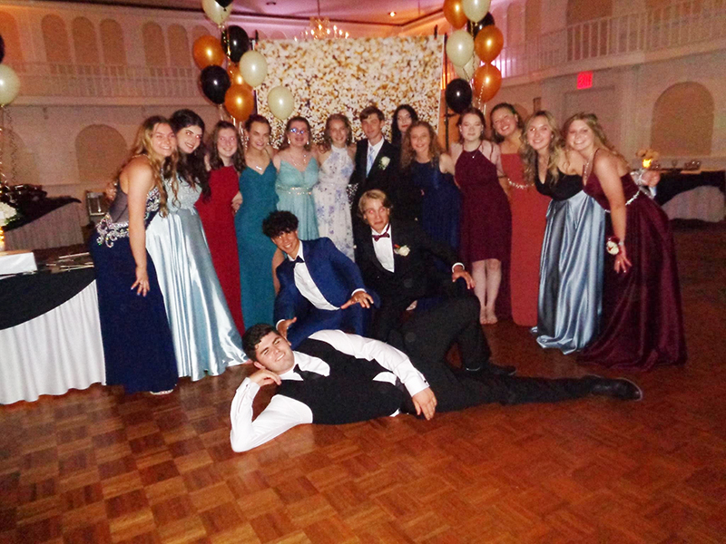 A large group of prom goers stand for the cmera. There is a young man laying on the floor in front of the group in his tuxedo.