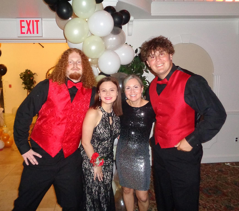 On either end is a young man wearing black shirt and pants and red vest and bow tie. in the center are two young women wearing black dresses. Behind them is an arch of black and white balloons.