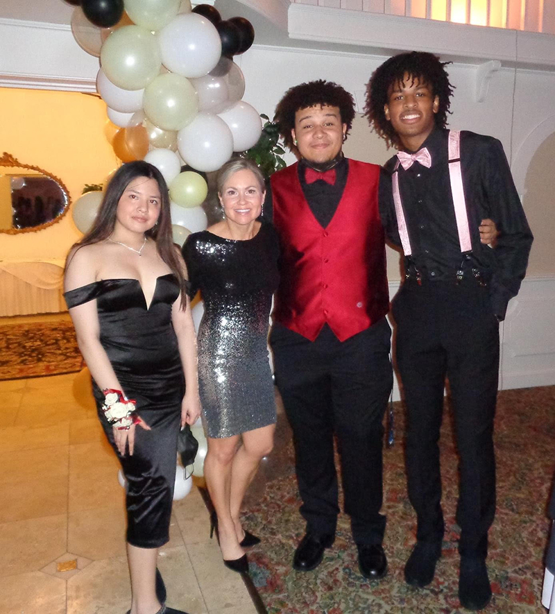 Three high school students stand with a teacher. The two boys on the right are wearing  black pants and shirts, one with a pink bow tie the other with a red bow tie and red vest. The girl on the left has a black off the shoulder dress on an the teacher, has a silver and black dress on.