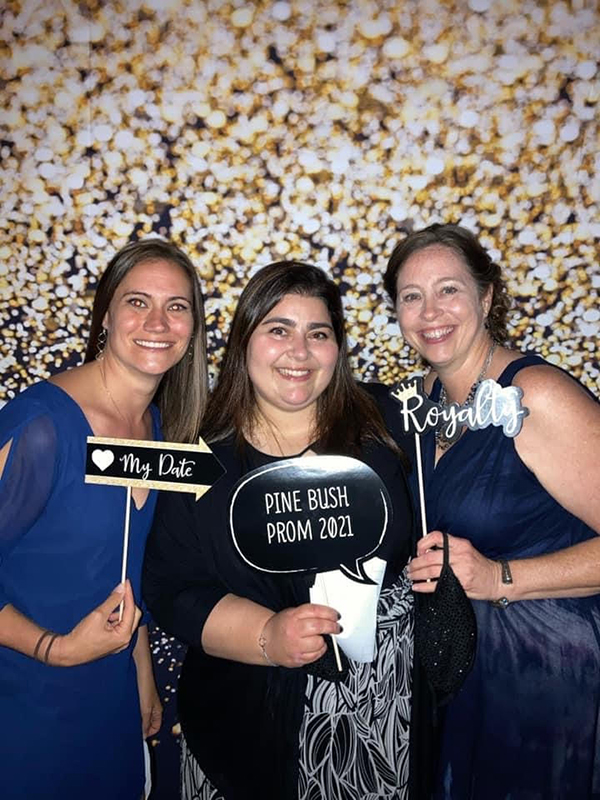 Three teachers all dressed in navy blue dresses, hold props that say "My Date" "Pine Bush Prom 2021" and "Royalty" 