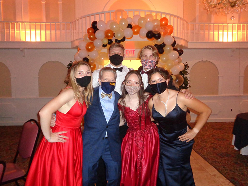 Three young women, two with red gowns, the other with a black gown, stand with two young men in the background and a man in a blue suit and plaid bow tie.