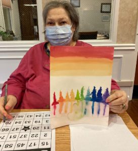 A woman in a red shirt wearing a mask holds up a painting with red and orange at the top and multicolor trees reflecting in water on the bottom.