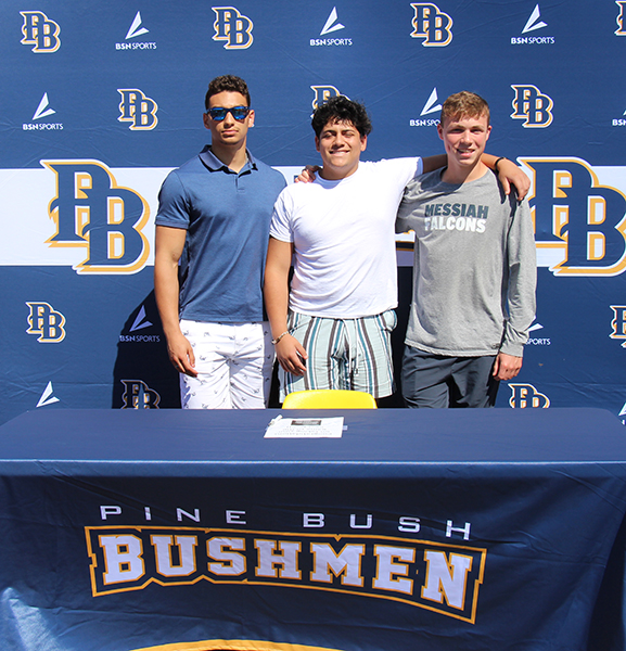 Three high school senior boys stand at a table with a blue tablecloth that says Pine Bush Bushmen. In the background says PB on a blue background.