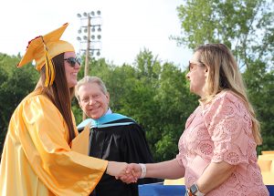A woman in a light pink dress shakes hands with a girl in a gold cap and gown with long brown hair.