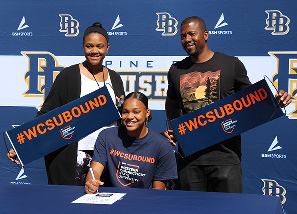 A young woman sits at a table signing a piece of paper. She is wearing a blue shirt that says Western  Connecticut. Behind her are a man and a woman holding banners that say #WCSU Bound