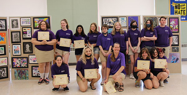 A group of  18 high school students, all wearing purple shirts and tan pants or shorts. They are in two rows, the front squating down. They are all holding certificates in front of them.
