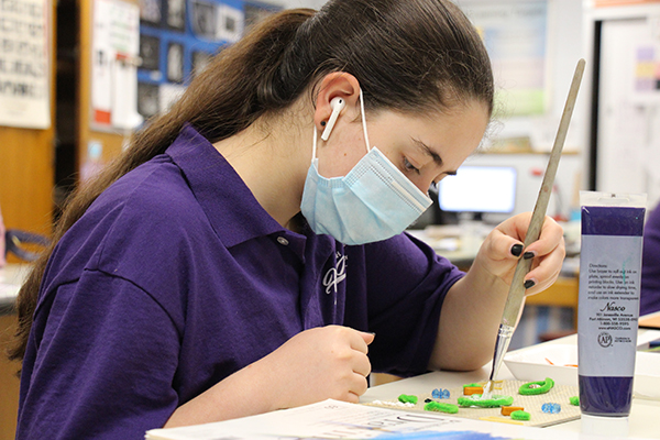 A high school student with long dark hair tied back in a poytail uses a paintbrugh to create a piece of art. She is wearing a blue face mask and earbuds.