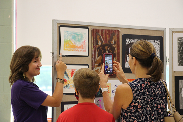 A high school student points at a piece of art on a wall and smiles as a woman takes her picture with a phone.