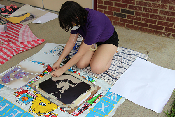 A female high school student dressed in a purple shirt and black shorts kneels  on the ground over a large piece of art she is working on.