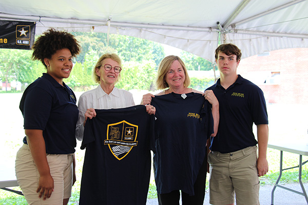 Two women in the center hold up blue shirts from the Leadership and Law Academy. There is a high school student on either side of them, smiling and both wearing khaki shorts and blue polo shirts.