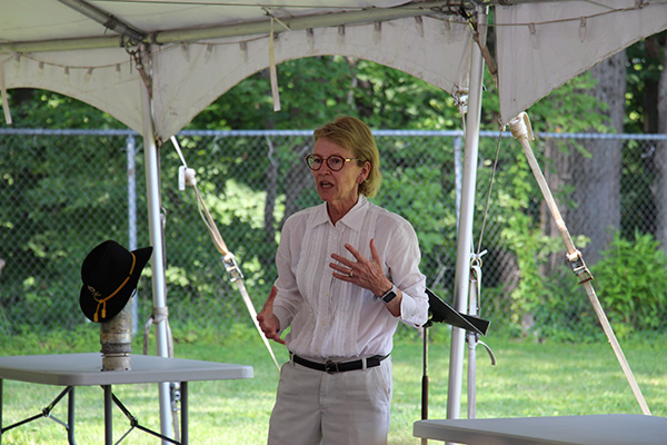 A woman with short blonde hair, wearing glasses, a white long sleeve shirt and tan pants talks, using her hands for emphasis.