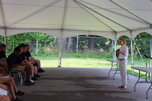 A woman with short blonde hair, wearing a white long sleeve shirt and tan pants stands in front of a group of high school students and talks.