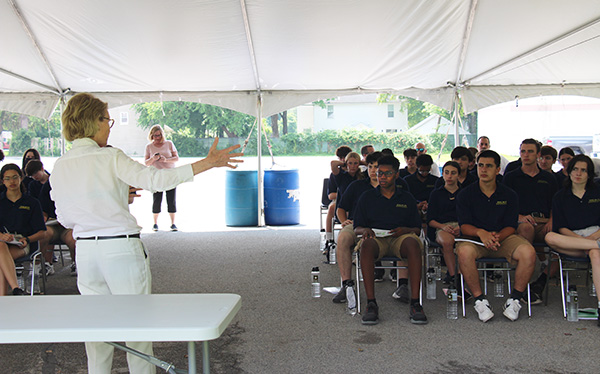 The back of a woman holding her arms out as she talks to a large group of high school students, sitting in two groups, under a large white tent. All of the students have navy blue shirts on and khaki shorts.