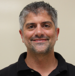 A man with salt and pepper hair and facial stubble wearing a black collared shirt smiles.