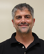 A man with salt and pepper hair and facial stubble wearing a black collared shirt smiles.